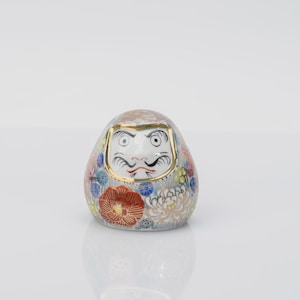 Peut inclure: Une figurine Daruma en céramique arrondie présentant un visage blanc avec des yeux, des sourcils et une moustache peints en noir. Le corps est orné d'un motif floral détaillé dans des tons de rouge, bleu, jaune et or, souligné par un liseré doré.