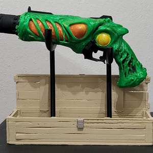 Peut inclure: Un pistolet factice vert et orange avec un design texturé unique, exposé sur un support noir dans un coffre en bois brun clair. Le pistolet présente un grand œil jaune et un canon noir.