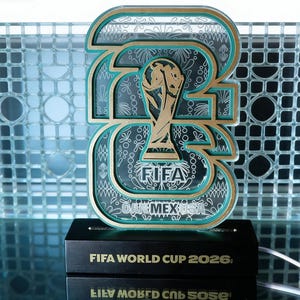 May include: A decorative FIFA World Cup 2026 sign. The sign features a gold trophy design within a teal and gold outline of the number 26. The base is black with the text "FIFA WORLD CUP 2026".