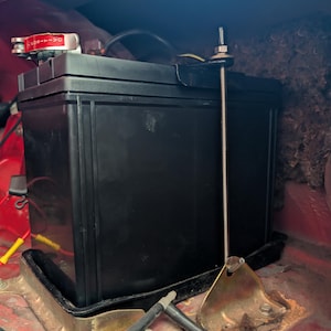 May include: A black car battery secured in a vehicle's compartment. The battery is held in place by a metal bracket and a threaded rod. The battery terminals have red and black connectors. The compartment has red paint.
