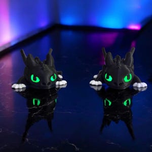 May include: Two black dragon figurines with glowing green eyes and white paws are displayed on a reflective surface. The dragons are 3D-printed and have a detailed, textured appearance. The background has a gradient of blue and purple.