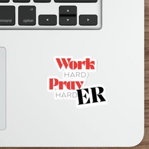 May include: A white sticker with the words "Work Hard Pray Harder" in red and black lettering. The sticker is on a white laptop.