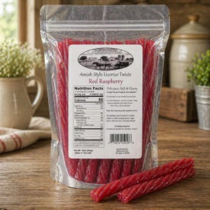 Op de afbeelding: Een doorzichtige plastic zak gevuld met rode zoethouttwists. Het etiket luidt "Amish Style Licorice Twists Red Raspberry" met voedingsinformatie. Twee zoethouttwists liggen buiten de zak op een houten oppervlak. De zak weegt 454g.
