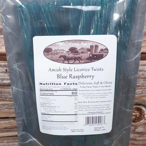 Op de afbeelding: Een transparante plastic zak gevuld met blauwe zoethouttwists. Het etiket luidt "Amish Style Licorice Twists Blue Raspberry" met een landelijke illustratie. Voedingsinformatie is zichtbaar. De zak bevat 227g zoethout.
