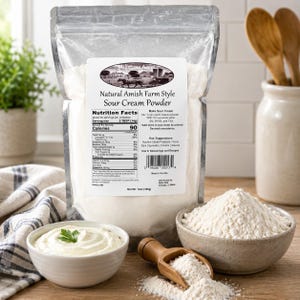 Op de afbeelding: Een doorzichtige plastic zak met Millstone Acres Natural Amish Farm Style zure roompoeder, nettogewicht 369g. Het etiket bevat een boerderijillustratie, voedingswaarden en instructies. Een kom met zure room en een kom met poeder staan ernaast.