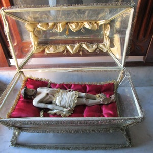May include: A religious artefact displayed in a glass case with gold trim. Inside, a figure lies on a red cushion, dressed in white cloth. The glass case has a hinged top. The figure has visible wounds.