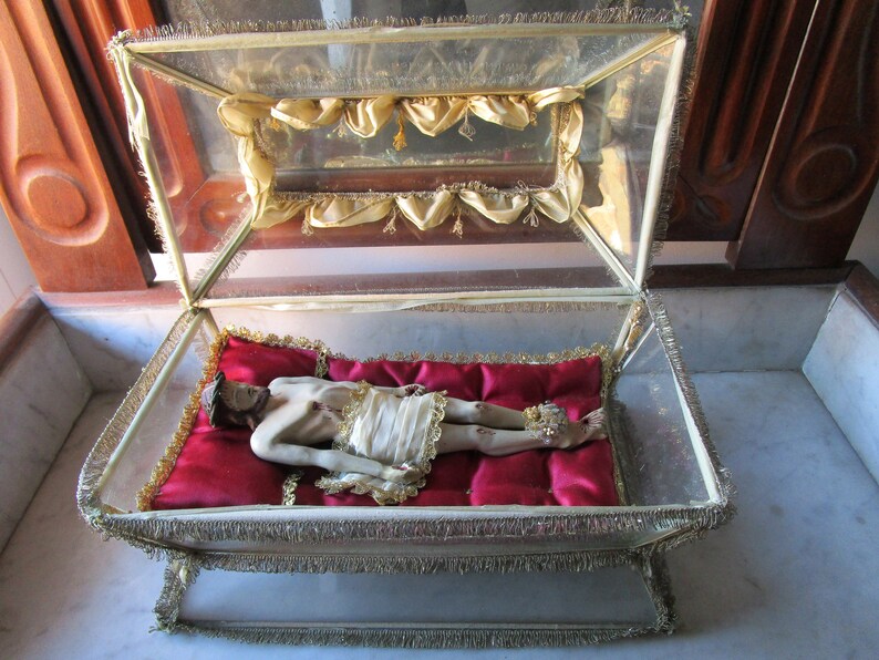 May include: A religious artefact displayed in a glass case with gold trim. Inside, a figure lies on a red cushion, dressed in white cloth. The glass case has a hinged top. The figure has visible wounds.