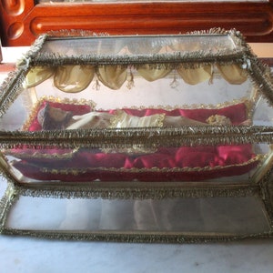 May include: A glass display case with gold trim, containing a figure dressed in white and gold, resting on a red satin pillow. The case is decorated with gold fringe. The case is approximately 25 cm long.