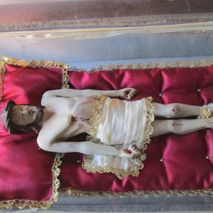May include: A religious figurine of a man with a beard and a crown of thorns, lying on a red cushion with gold trim. The figure wears a white cloth and has decorative ankle embellishments.