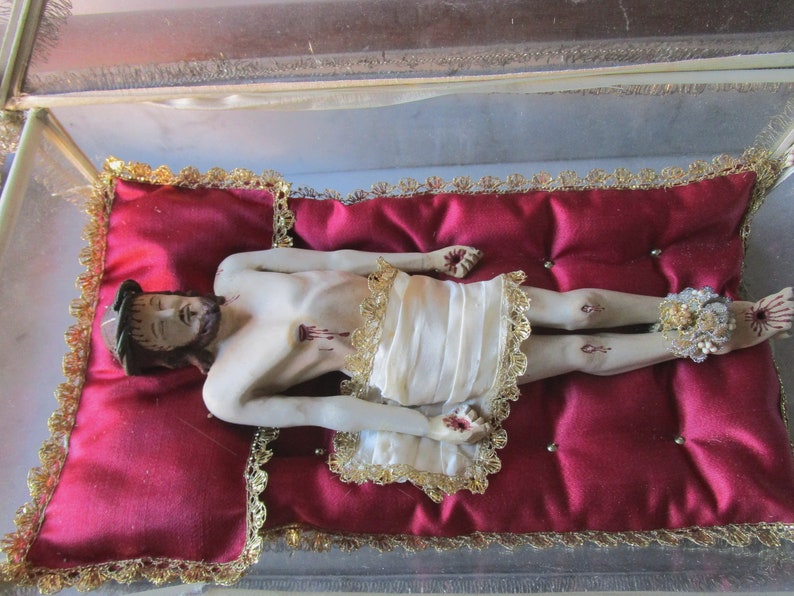 May include: A religious figurine of a man with a beard and a crown of thorns, lying on a red cushion with gold trim. The figure wears a white cloth and has decorative ankle embellishments.
