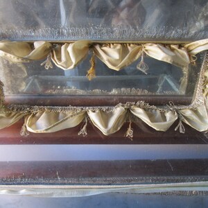 May include: A vintage display case with a glass front and a wooden frame. The case is decorated with cream-coloured fabric valances and decorative fringe. The glass appears to be slightly aged, adding to its antique charm.