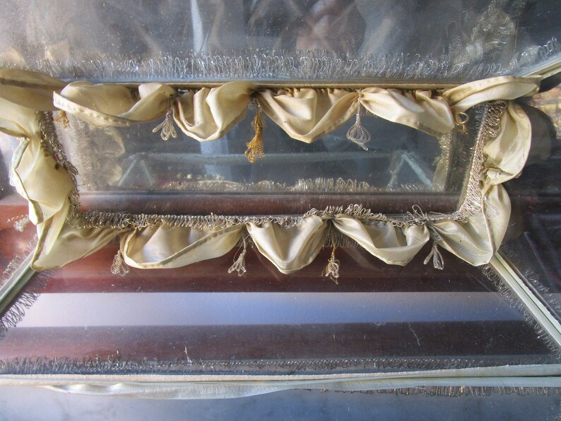 May include: A vintage display case with a glass front and a wooden frame. The case is decorated with cream-coloured fabric valances and decorative fringe. The glass appears to be slightly aged, adding to its antique charm.