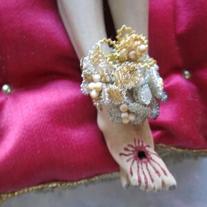 May include: A detailed depiction of a foot with a wound, adorned with a decorative ankle bracelet. The bracelet features gold and silver wire with pearl-like accents. The foot rests on a red velvet cushion.