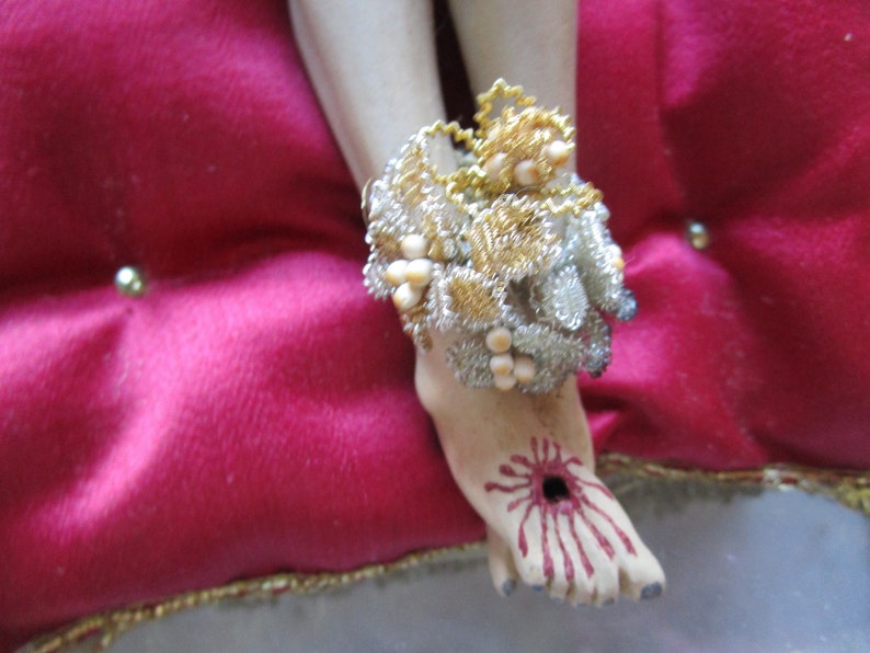May include: A detailed depiction of a foot with a wound, adorned with a decorative ankle bracelet. The bracelet features gold and silver wire with pearl-like accents. The foot rests on a red velvet cushion.
