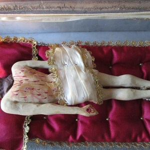 May include: A religious figurine of a figure lying on a red velvet cushion. The figure has painted wounds and is covered with a white cloth with gold trim. The cushion is framed with gold trim.