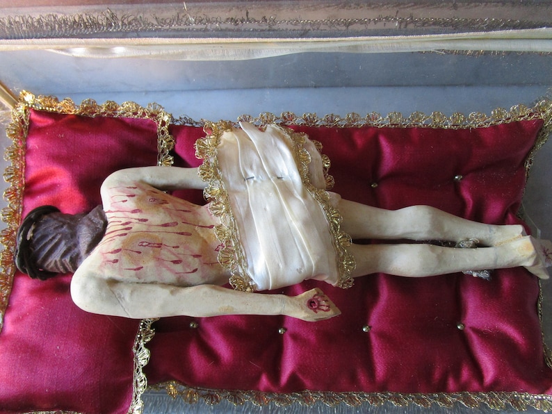May include: A religious figurine of a figure lying on a red velvet cushion. The figure has painted wounds and is covered with a white cloth with gold trim. The cushion is framed with gold trim.
