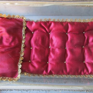 May include: A small, rectangular, red satin cushion with a quilted design and gold trim. The cushion is adorned with small, gold-coloured beads and is set inside a clear, rectangular box with gold trim.