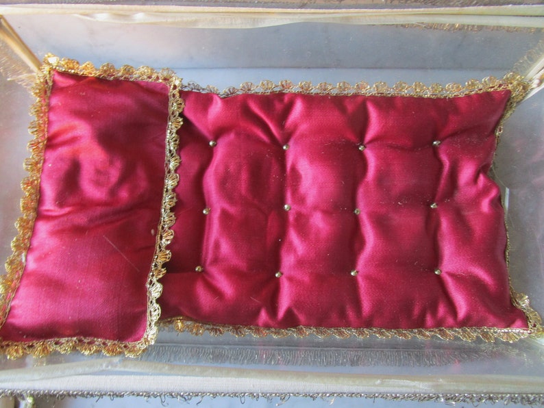 May include: A small, rectangular, red satin cushion with a quilted design and gold trim. The cushion is adorned with small, gold-coloured beads and is set inside a clear, rectangular box with gold trim.