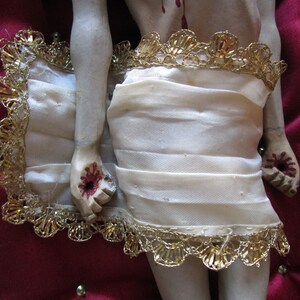 May include: A religious figurine with a cream-coloured cloth wrapped around the waist, edged with gold trim. The hands and feet show painted wounds. It rests on a red velvet cushion with gold trim. A detailed depiction.