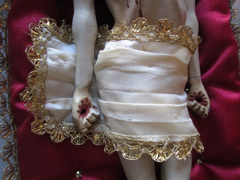 May include: A religious figurine with a cream-coloured cloth wrapped around the waist, edged with gold trim. The hands and feet show painted wounds. It rests on a red velvet cushion with gold trim. A detailed depiction.