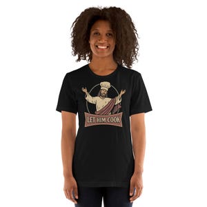 May include: Black t-shirt featuring a graphic of a figure in a chef's hat and robe, with arms outstretched. The design is encircled by a brown banner with the text "LET HIM COOK". The shirt is made of a soft material.