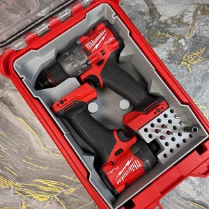 May include: Red and black Milwaukee FUEL power tools set in a red hard case. The set includes a drill and a smaller tool, along with a bit holder containing various drill bits. The case is open, revealing the tools neatly organized inside.