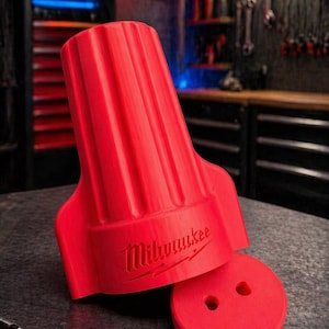 May include: A red Milwaukee wire connector with a ribbed top and a separate circular piece with two holes. The Milwaukee logo is visible on the connector. The background shows a workshop setting with tools and a neon sign.