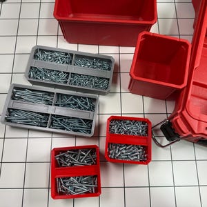 以下が含まれることがあります： さまざまなネジと収納容器。 グレーのプラスチック製オーガナイザーにはネジが詰め込まれ、赤いプラスチック製のビンと大きな赤いツールボックスが並んでいます。 アイテムは白いタイルの表面に配置されています。