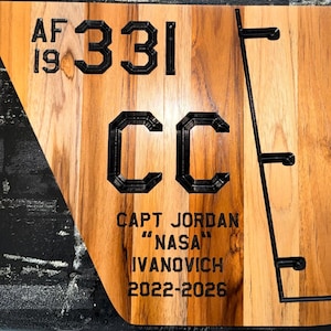 May include: Wooden plaque with engraved black text and design elements. The text includes "AF 19", "331", "CC", "CAPT JORDAN", "NASA", "IVANOVICH", and "2022-2026". The plaque has a natural wood grain finish and a black border.