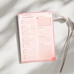 May include: A pink weekly planner with floral accents and the German text "Undatierter Wochenplaner." The planner includes sections for daily tasks, priorities, and notes. The bottom of the planner has the text "ES GEHT UM FORTSCHRITT, NICHT UM PERFEKTION."