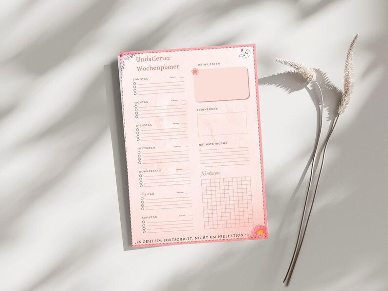 May include: A pink weekly planner with floral accents and the German text "Undatierter Wochenplaner." The planner includes sections for daily tasks, priorities, and notes. The bottom of the planner has the text "ES GEHT UM FORTSCHRITT, NICHT UM PERFEKTION."