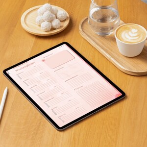 May include: A digital tablet displaying a pink weekly planner with the German text "Wochenplaner". A white stylus rests beside the tablet. A cup of coffee, a glass of water, and a plate of white confections sit on a wooden table.