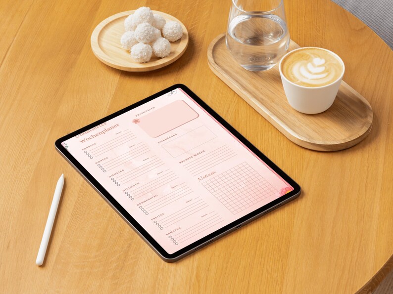 May include: A digital tablet displaying a pink weekly planner with the German text "Wochenplaner". A white stylus rests beside the tablet. A cup of coffee, a glass of water, and a plate of white confections sit on a wooden table.