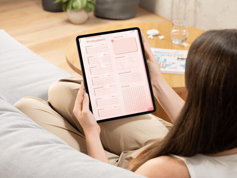 May include: A digital tablet displaying a pink and white weekly planner with the German title "Wochenplaner". The tablet is held by a person, with the screen facing the viewer. The planner includes sections for daily tasks and notes.