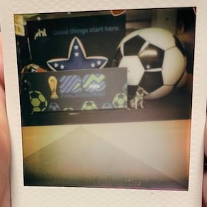 May include: A square instant photograph featuring a display with a large black and white soccer ball, a blue star with white stars, and a banner reading PHL Good things start here and FIFA Philadelphia with a gold trophy icon and soccer ball graphics.