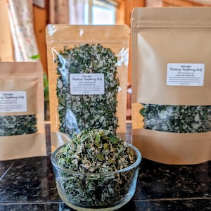 May include: Three brown kraft paper pouches of varying sizes containing dried Blackcap Raspberry Leaf herbal tea, each with a white label. A clear glass bowl filled with the same dried green and white leaf tea sits in the foreground.