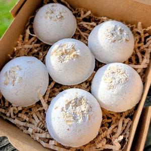 May include: Six round, off-white bath bombs arranged in a cardboard box. Each spherical bath bomb features a textured surface with a cluster of dried oat flakes embedded on the top. The items are nestled within brown shredded paper filler.