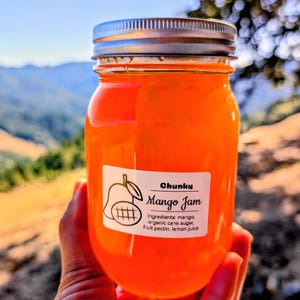 May include: A jar of chunky mango jam with a silver lid. The label reads "Chunky Mango Jam" with a mango illustration and ingredient list. The jam is a vibrant orange color, and the jar is held up against a scenic outdoor background.