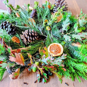 May include: A decorative wreath arrangement featuring green pine and cedar branches, brown pinecones, dried orange slices, small green acorns, and dried oak leaves. The composition is layered with natural textures and earthy tones, creating a dense, circular display of seasonal botanical elements.