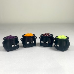 May include: Four small black cauldrons arranged in a row, each featuring a simple white face with two circular eyes and a curved mouth. Each cauldron is filled with a different colored substance: purple, orange, dark red, and bright lime green.