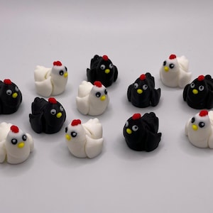 Può includere: Una collezione di piccole figurine decorative a forma di pollo. Le figurine sono in due colori, nero e bianco, con creste rosse e becchi gialli. I polli sono disposti su una superficie bianca.