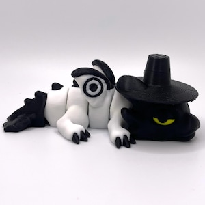 May include: A whimsical, stylized dragon figurine in black and white, with a black hat. The dragon is lying down, with its body segmented in white and black. The dragon's eyes are yellow.