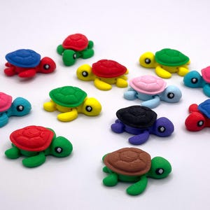May include: A collection of colorful miniature turtle figurines. Each turtle has a different colored shell, including red, blue, green, pink, and brown. The turtles are arranged on a white surface.