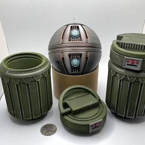May include: A collection of olive green and metallic gray 3D-printed objects. The objects include two cylindrical containers, a spherical object, and a circular object with a handle. The spherical object has turquoise accents. A US quarter is included for scale.