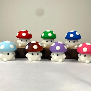 May include: A collection of seven small mushroom figurines with white bodies and caps in various colors including red, green, blue, light blue, brown, purple, and pink. Each mushroom features white spots on the cap and small black eyes.