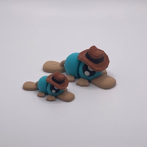 May include: Two whimsical, handmade figurines. One large and one small, both feature a blue body, brown cowboy hat, and tan feet. The figures are lying down on a white surface. The larger figure is approximately 7.6 cm long.