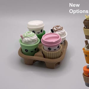 May include: A set of four miniature beverage cups with smiling faces in a brown cardboard carrier, plus three additional cup options on the right labeled New Options. The cups feature lids, whipped cream toppings, and textured sleeves.