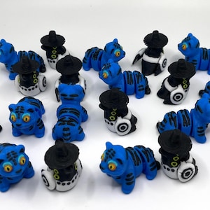 Op de afbeelding: Een verzameling kleine, blauwe tijgerbeeldjes met zwarte strepen en gele ogen. Sommige beeldjes dragen zwarte hoeden en hebben witte en zwarte accenten. De beeldjes zijn gerangschikt op een wit oppervlak.