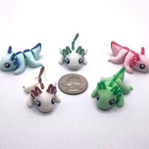 Può includere: Cinque figurine di axolotl colorati in azzurro, bianco, rosa e verde, con dettagli a contrasto sulle branchie e sulle pinne. Una moneta è inclusa come riferimento delle dimensioni. Gli axolotl sono su una superficie bianca.