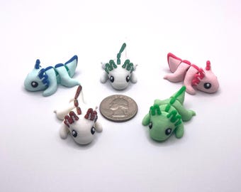 May include: Five colorful axolotl figurines in light blue, white, pink, and green, with contrasting gill and fin details. A US quarter is included for size reference. The axolotls are lying down on a white surface.